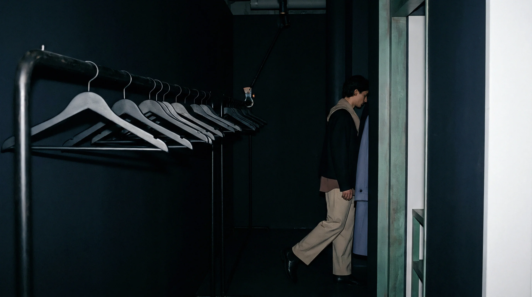 Rows of wooden hangers rest on a metal clothing rack within a dark interior beside a standing figure and an entrance.
