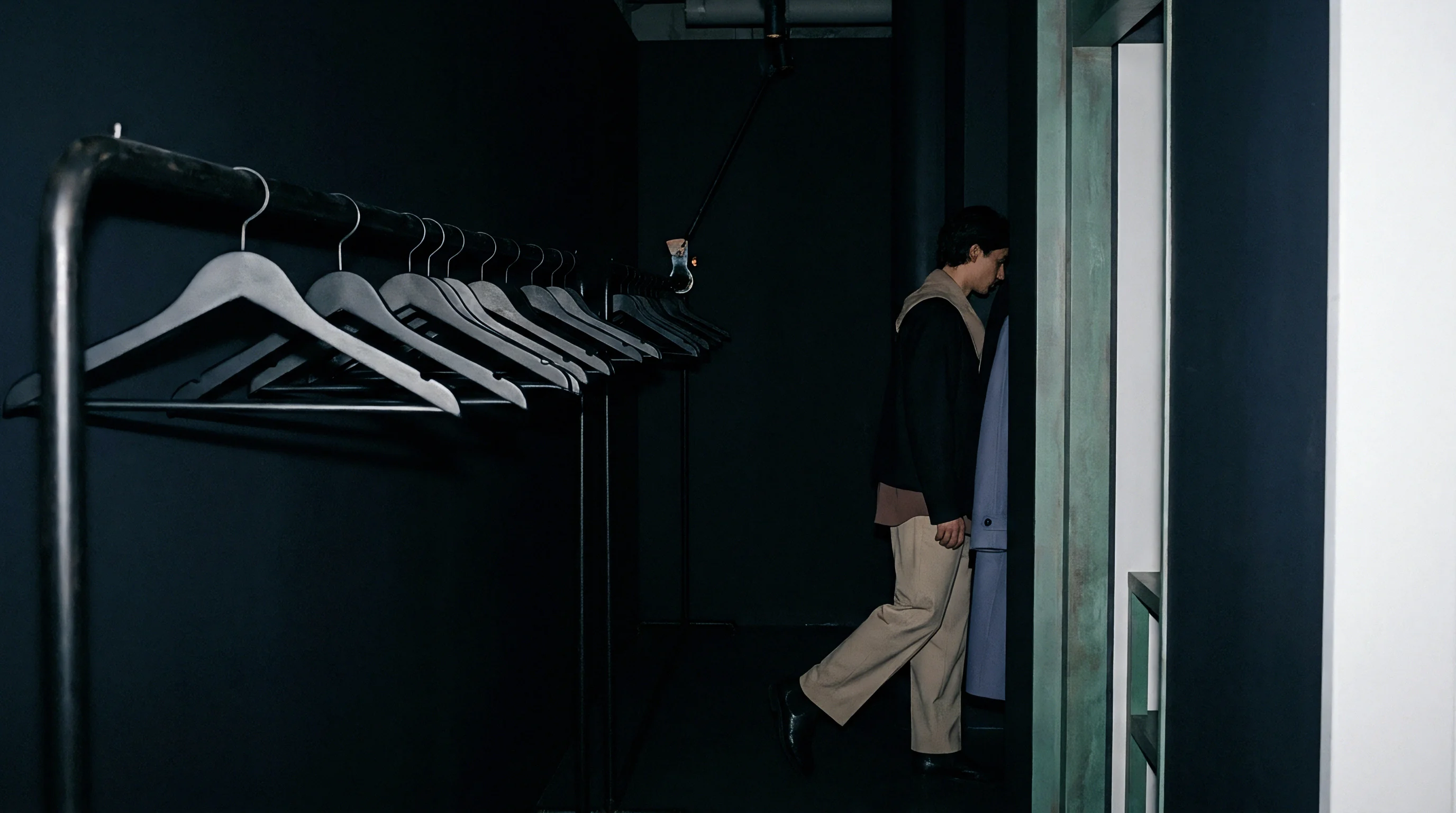 Rows of wooden hangers rest on a metal clothing rack within a dark interior beside a standing figure and an entrance.