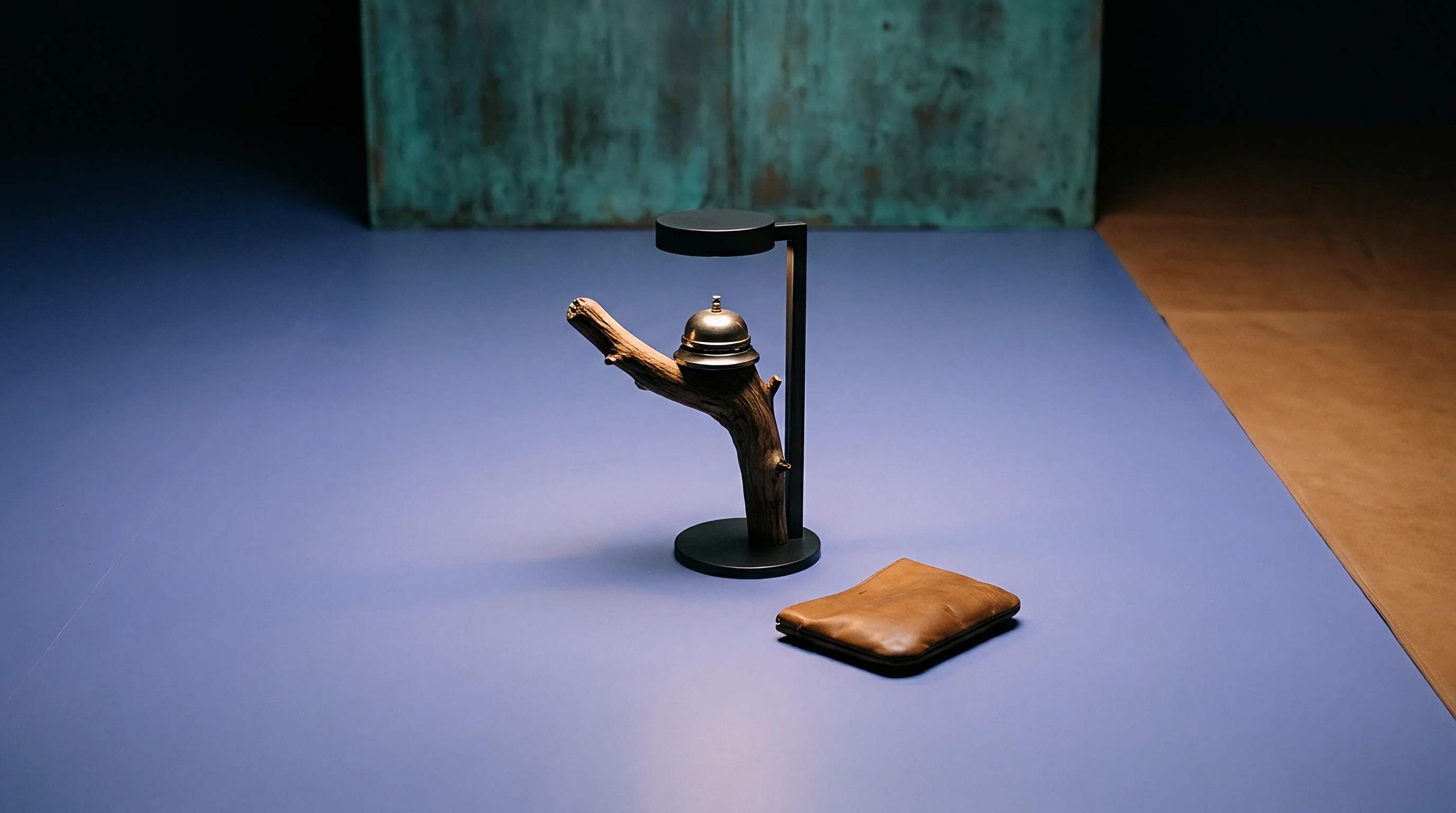 A wooden branch lamp holding a metallic service bell sits beside a leather wallet on a flat blue manufacturing floor surface.