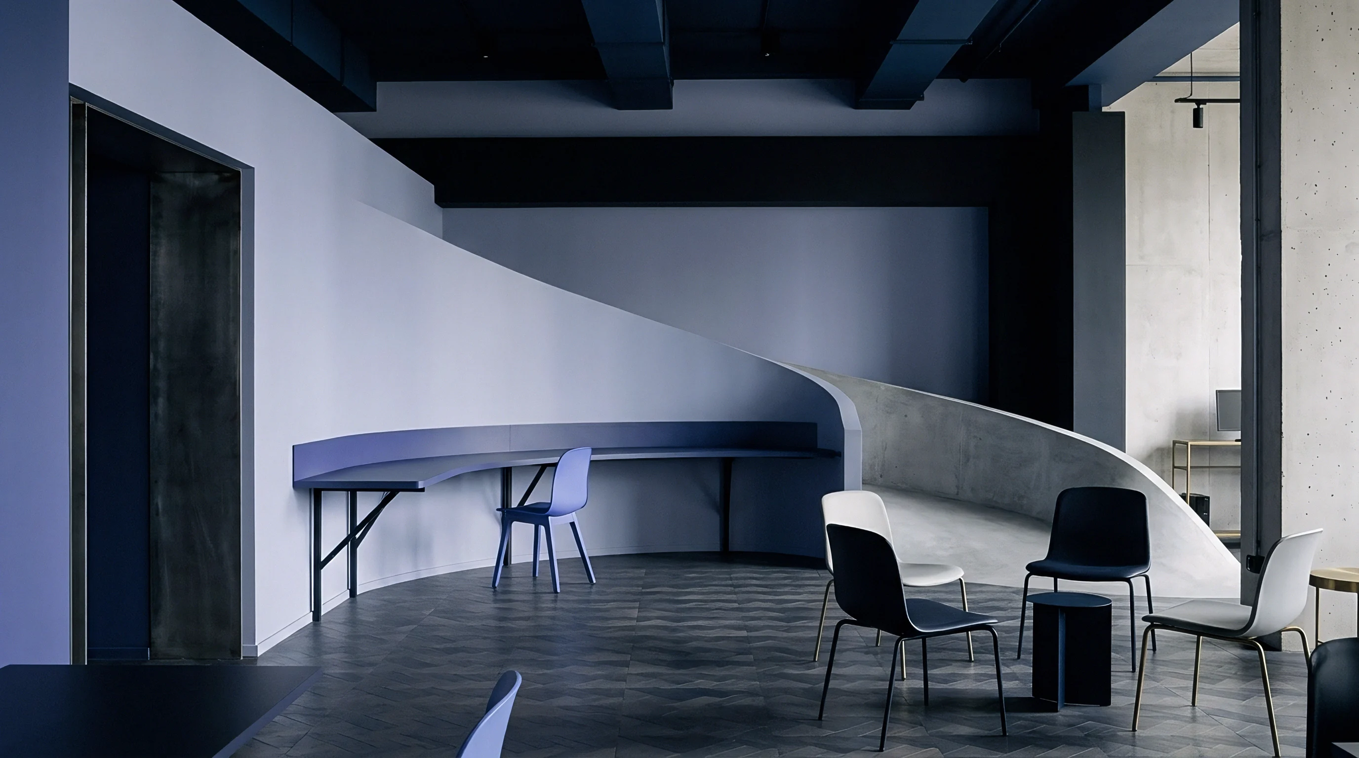 An industrial design studio features a sweeping concrete ramp, a contoured perimeter desk, and grouped chairs on a dark wood floor.