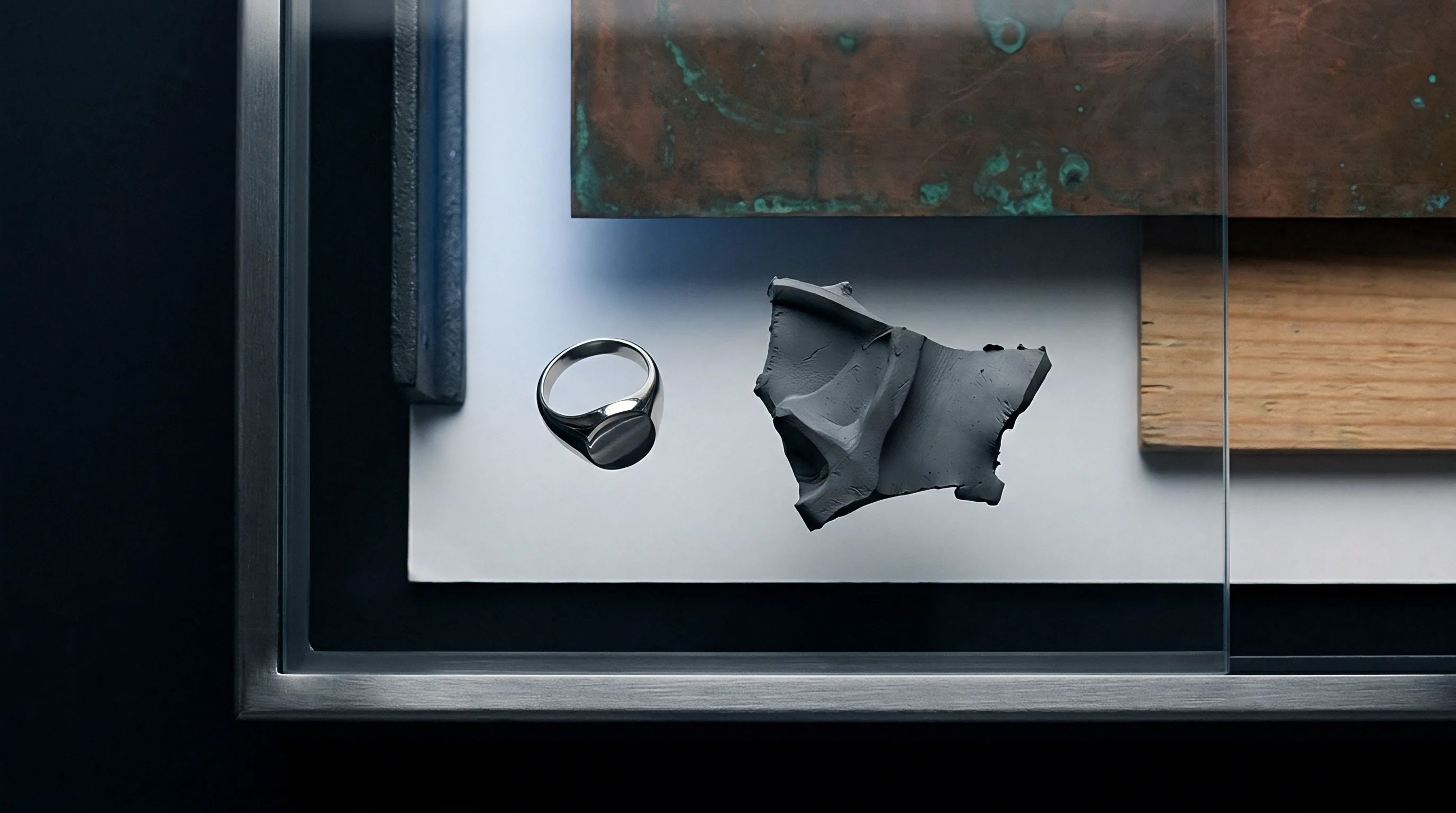 A polished metal ring sits beside an amorphous grey clay fragment on white paper framed with oxidized copper and timber planks.