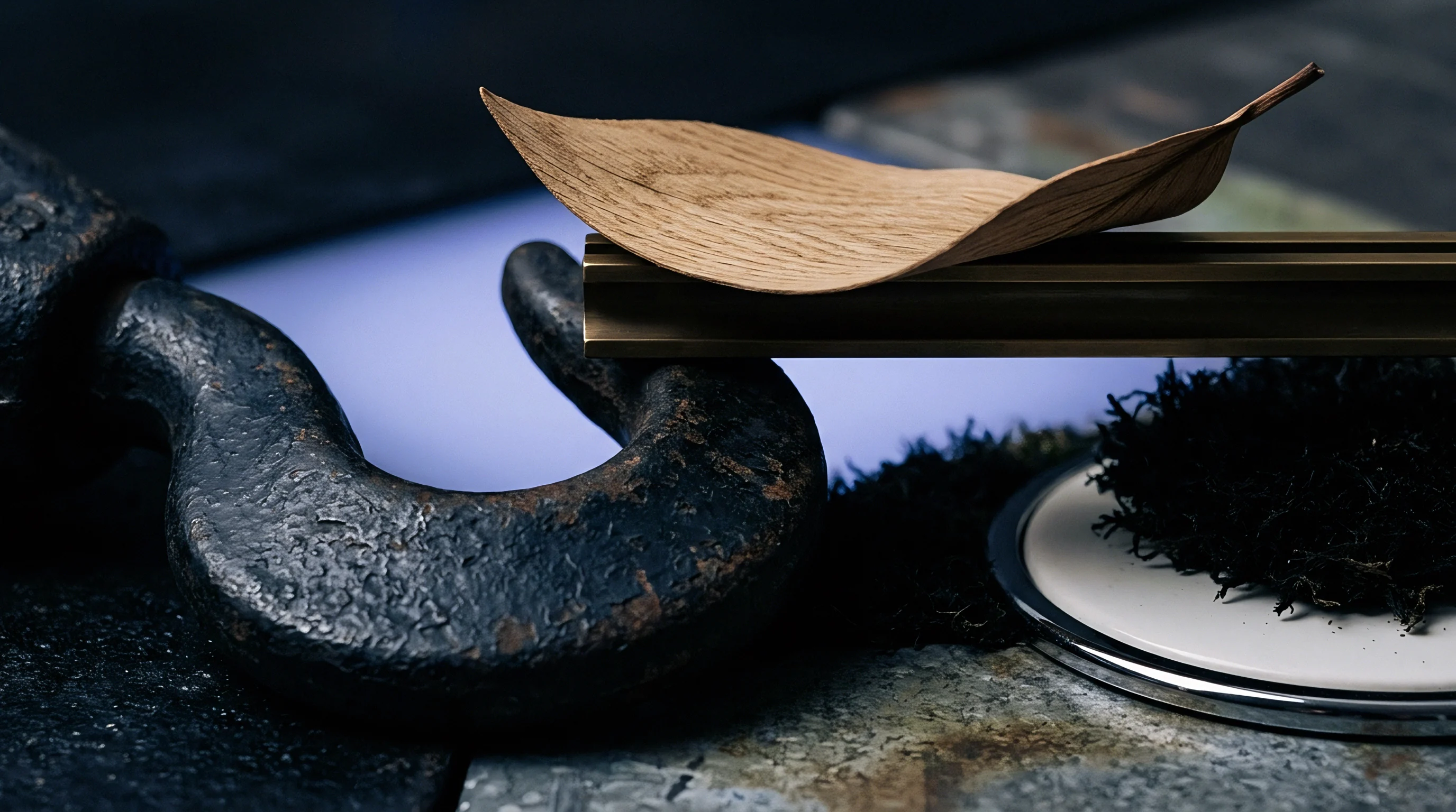 A textured cast iron hook provides a heavy industrial base for a brass bar supporting a light wooden leaf shape.