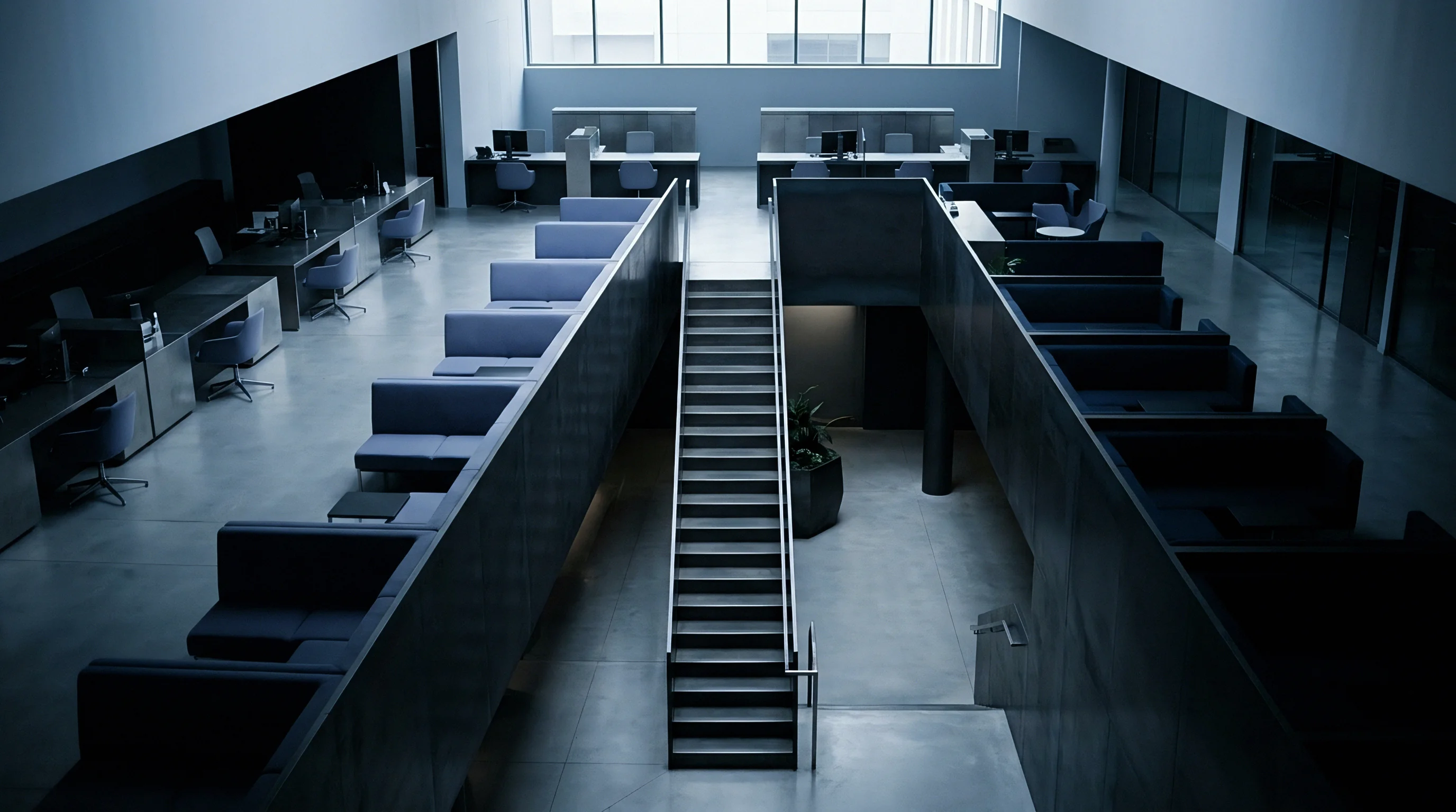 Rows of modular workstations line the dual level office floor divided by a central staircase leading towards large upper windows.