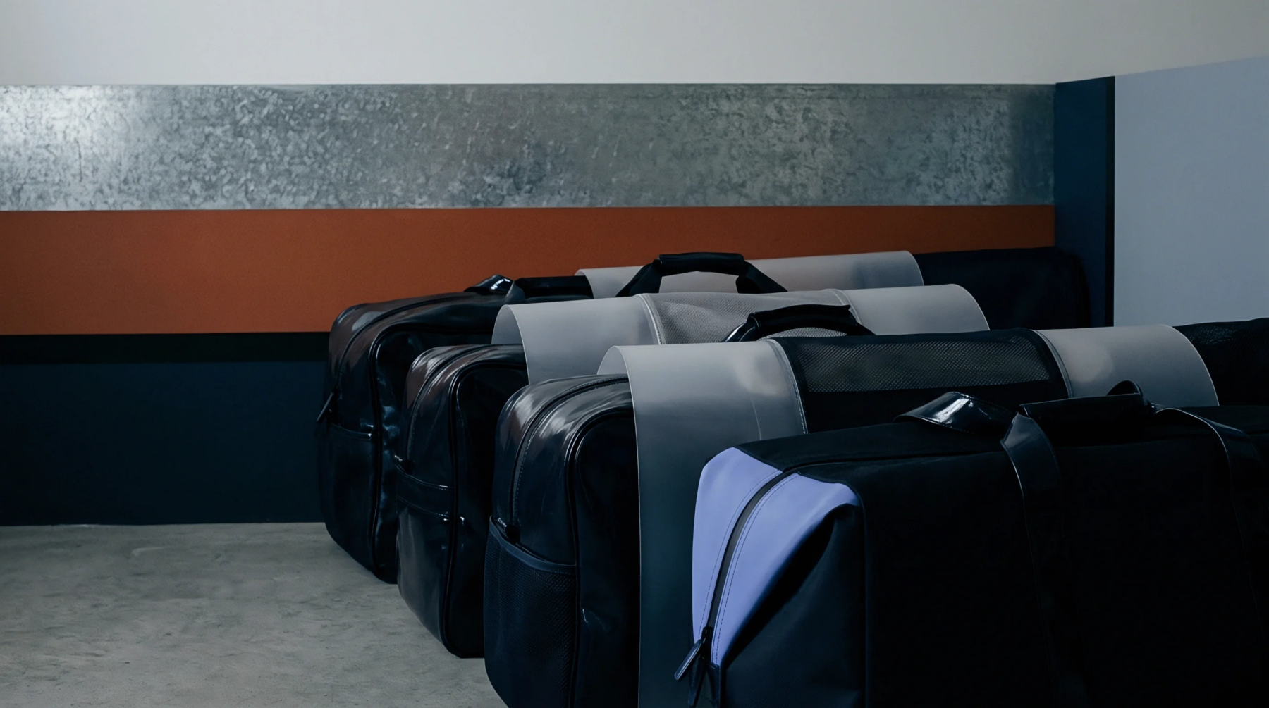 Black fabric transit bags with transparent protective films stand aligned sequentially along an industrial workshop floor beneath metal wall panels.